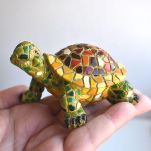 Puede incluir: Una pequeña figurita de tortuga de mosaico de colores. El caparazón está hecho de pequeñas baldosas de colores en tonos rojos, naranjas, amarillos, verdes y marrones. La tortuga está de pie sobre sus cuatro patas, con la cabeza levantada.