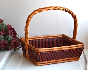 May include: A woven wicker basket with a brown handle and a red and black striped lining. The basket is empty.