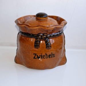 Puede incluir: Un tarro de cerámica marrón con tapa, diseñado para parecerse a un saco. El tarro tiene un detalle de cuerda marrón oscuro alrededor del centro y la palabra "Zwiebeln" está impresa en la parte delantera. La tapa tiene un pequeño pomo marrón oscuro.