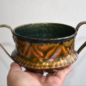 Può includere: Una ciotola decorativa in ceramica con due manici. Presenta una superficie strutturata e smaltata con sfumature di arancione, verde e blu. L'interno è verde scuro. I manici sono metallici.