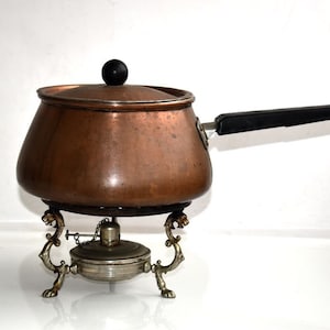 Puede incluir: Olla de fondue de cobre con mango negro y soporte plateado con patas decorativas. La olla tiene una tapa con un pomo negro.
