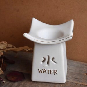 Può includere: Bruciatore di incenso in ceramica bianca con base quadrata e piattaforma rialzata. Il bruciatore ha un bordo dorato e la parola "WATER" scritta sul davanti in inglese e caratteri cinesi.