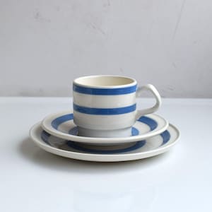May include: A ceramic cup and saucer set featuring a white base with bold, horizontal blue stripes. The cup has a curved handle and a stepped base, while the matching saucer displays a concentric blue striped pattern on its flat surface.