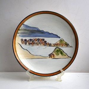 May include: A white ceramic plate with a hand-painted scene of a coastal town with houses, hills, and a blue sky. The plate has a brown and green rim.