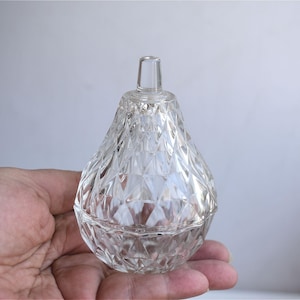 Puede incluir: Un tarro de cristal transparente facetado con tapa, sostenido en una mano. El tarro tiene forma de lágrima con un patrón de diamantes. La tapa tiene un pequeño asa cilíndrica. El tarro es probablemente para guardar objetos pequeños o como pieza decorativa.