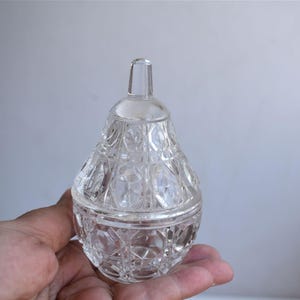 Könnte beinhalten: Ein klares, antikes Glasgefäß mit Deckel. Das Gefäß hat ein strukturiertes, geometrisches Muster und einen kleinen Griff am Deckel. Das Gefäß wird in der Hand einer Person gehalten.