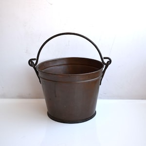 Peut inclure: Seau en cuivre vintage avec une finition marron foncé. Le seau a un corps arrondi et une poignée arquée robuste en métal foncé. La poignée est fixée au seau avec des supports incurvés. Le seau repose sur une base sombre.