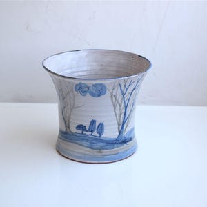 Pode incluir: Um vaso de cerâmica com borda alargada, pintado com uma cena de paisagem em azul e cinza. O design apresenta árvores estilizadas e uma linha do horizonte. O vaso tem um fundo branco e cinza.