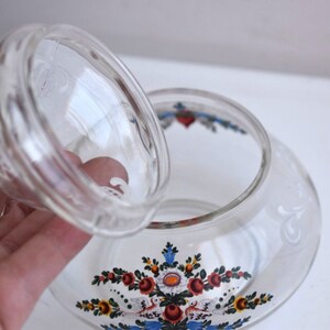K&ouml;nnte beinhalten: Eine klare Glasdose mit Deckel, verziert mit einem Blumendesign in Rot, Blau, Gelb und Wei&szlig;. Die Dose hat eine runde Form und eine breite &Ouml;ffnung.