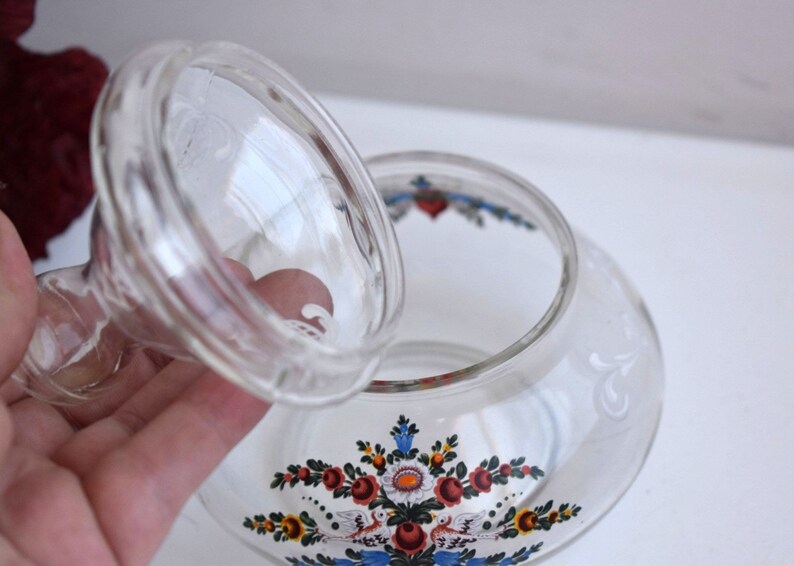 K&ouml;nnte beinhalten: Eine klare Glasdose mit Deckel, verziert mit einem Blumendesign in Rot, Blau, Gelb und Wei&szlig;. Die Dose hat eine runde Form und eine breite &Ouml;ffnung.