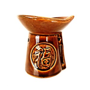 Può includere: Un bruciatore per olio in ceramica marrone con una parte superiore larga e poco profonda e una base rotonda. Il bruciatore presenta un design circolare in rilievo con il carattere cinese per "fortuna" al centro.