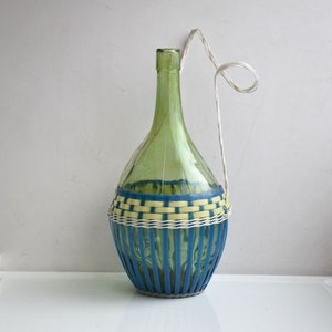 Könnte beinhalten: Eine grüne Glasflasche mit einem blauen und weißen geflochtenen Korb drumherum. Die Flasche hat einen Seilgriff.