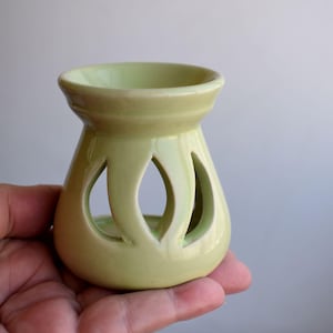 Può includere: Un diffusore di oli essenziali in ceramica verde chiaro. Il diffusore ha una base rotonda, un corpo bulboso con tre ritagli decorativi e una piccola ciotola nella parte superiore. Il diffusore di oli è progettato per contenere oli essenziali e rilasciare la loro fragranza.