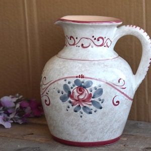 Puede incluir: Una jarra de cerámica blanca con un borde y un asa rojos. La jarra está decorada con un diseño floral en tonos rosa, azul y verde.