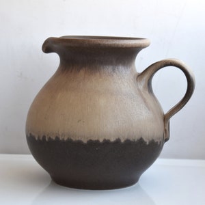 Può includere: Un'anfora in ceramica marrone e beige con corpo arrotondato e manico. L'anfora ha una finitura smaltata e una fascia marrone intorno alla base.