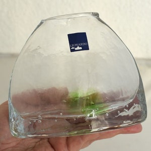 Może przedstawiać: Przezroczysty szklany wazon o zaokrąglonym, asymetrycznym kształcie. Wazon ma teksturowaną powierzchnię i małą niebieską etykietę z napisem "LEONARDO". W szkle widoczne są pęcherzyki powietrza, a wewnątrz zielone i różowe odcienie.