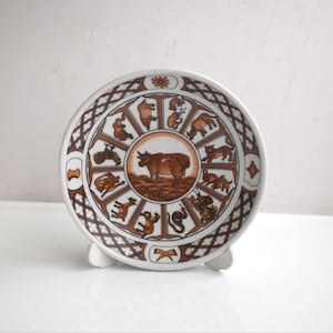 Può includere: Un piatto rotondo in ceramica con un design zodiacale marrone e bianco. Il centro del piatto raffigura una mucca marrone, circondata da simboli zodiacali in un motivo circolare. Il piatto è sostenuto da un supporto bianco.