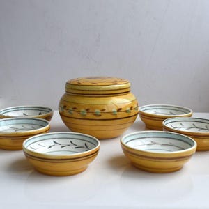 Könnte beinhalten: Ein Set aus sieben gelben Keramikschalen mit einem blauen und braunen Blumendesign. Die Schalen sind in einem Kreis um eine größere gelbe Keramikdose mit Deckel angeordnet. Die Dose hat ebenfalls ein blaues und braunes Blumendesign.