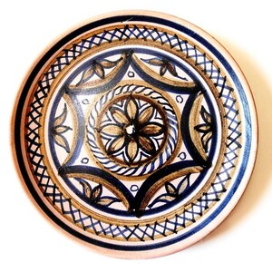 May include: A round ceramic plate with a detailed, hand-painted design. The plate features a central floral motif in brown and black, surrounded by blue and brown geometric patterns. The rim is a light pink color.