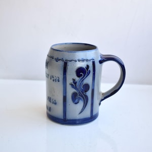 May include: A gray ceramic beer stein with blue and white floral designs. The stein has the text "Est 1927" and "Pils" on the front.