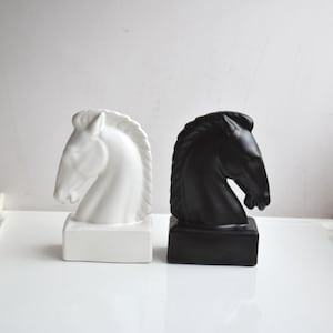 Puede incluir: Dos esculturas de cabeza de caballo de cerámica, una blanca y otra negra, sobre una superficie blanca.