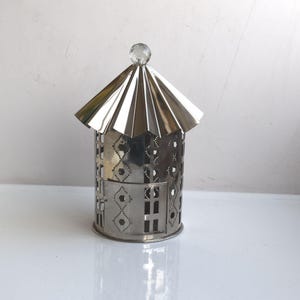 Può includere: Un portacandele in metallo color argento a forma di casa. La casa ha un tetto a punta e una sfera di cristallo trasparente sulla parte superiore. I lati sono decorati con motivi ritagliati e forme geometriche.