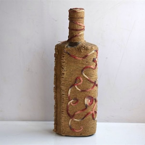Puede incluir: Una botella de vidrio envuelta en cuerda natural con acentos rojos y blancos. La cuerda está tejida en un patrón decorativo.