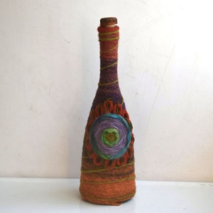 Puede incluir: Una botella de vidrio envuelta en hilo de colores con un diseño floral decorativo en tonos verdes, morados y azules.