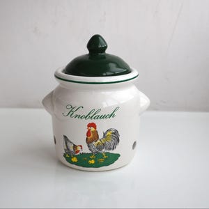 Può includere: Contenitore per aglio in ceramica bianca con coperchio e manico verde scuro. Il contenitore presenta un'illustrazione di gallo e gallina con la scritta "Knoblauch" in corsivo. Il contenitore ha piccoli fori per la ventilazione.