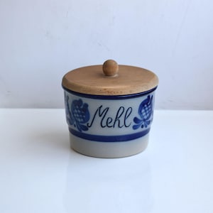 Könnte beinhalten: Eine blaue und weiße Keramikdose mit einem Holzdeckel. Die Dose hat ein blaues und weißes Blumenmuster und das Wort "Mehl" steht auf der Vorderseite.