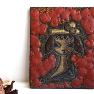 Könnte beinhalten: Eine Keramik-Wandfliese mit einem roten, braunen und goldenen Design. Die Fliese zeigt ein stilisiertes Bild einer Person, die einen Hut mit einem Blumendesign trägt.