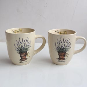 Puede incluir: Dos tazas de cerámica color crema con un diseño de planta de lavanda. Cada taza presenta una maceta marrón con una planta de lavanda y la palabra "Lavender" debajo. Una pequeña etiqueta con la palabra "Lavender" cuelga del borde.