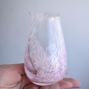 Puede incluir: Un jarrón de vidrio transparente con la parte superior acampanada y un cuerpo redondeado. El jarrón presenta un delicado patrón moteado rosa y blanco, con vetas blancas plumosas. El jarrón se sostiene en una mano.