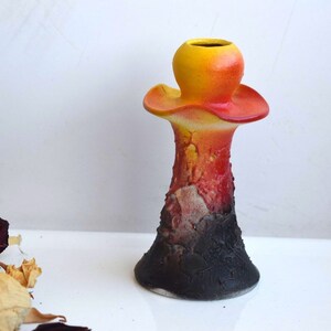 May include: A ceramic candle holder with a textured, black base and a yellow, orange, and red top. The top is shaped like a mushroom with a wide, flat brim.