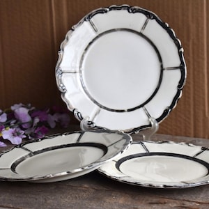 May include: Three white ceramic plates with black and silver trim. The plates have a scalloped edge and a decorative design.