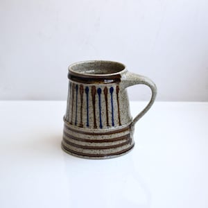 May include: A brown and gray ceramic mug with a blue and brown striped design. The mug has a handle and a speckled glaze.