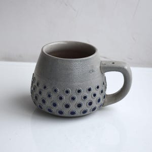 Op de afbeelding: Grijze keramische mok met een afgeronde body en een handvat. De mok is versierd met een patroon van donkerblauwe cirkels. De mok heeft een getextureerd oppervlak en een eenvoudig ontwerp.