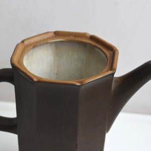 Może przedstawiać: Brązowy ceramiczny czajnik o ośmiokątnym kształcie z brązowym uchwytem. Czajnik ma jasnobrązową krawędź i białe wnętrze.