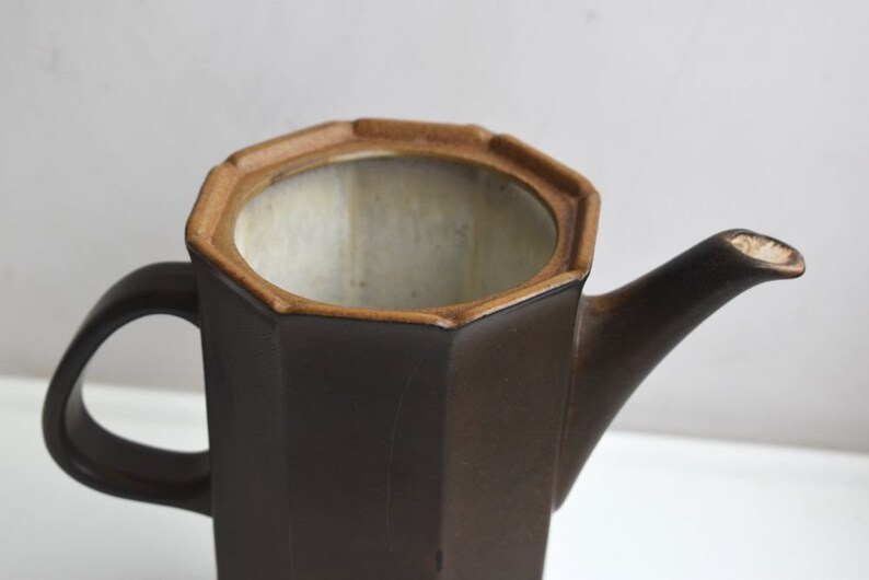 Może przedstawiać: Brązowy ceramiczny czajnik o ośmiokątnym kształcie z brązowym uchwytem. Czajnik ma jasnobrązową krawędź i białe wnętrze.