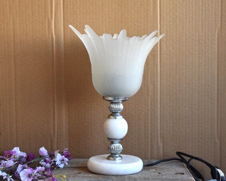 May include: A white table lamp with a frosted glass shade shaped like a flower. The lamp has a silver base with a white marble base.