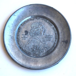 May include: A silver-coloured metal plate with a raised image of a man in a crown and a coat of arms. The plate is inscribed with the text "WERNER REICHGRAF VON INNOHEIM" and "GEBOREN IN WERNSTEIN AM 16. APRIL 1628" and "GESTORBEN IN INNOHEIM AM 26. APRIL 1693".
