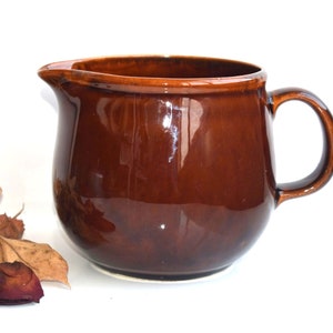Peut inclure: Une cruche en céramique marron avec une anse et un bec verseur. La cruche a une finition brillante et une forme arrondie. Des feuilles et des pétales séchés sont éparpillés près de la cruche.