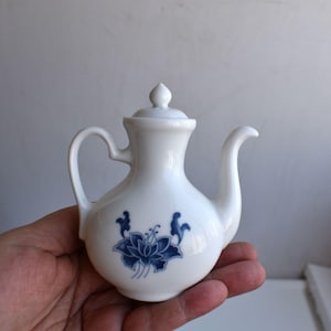 Könnte beinhalten: Kleine, weiße Keramik-Teekanne mit Deckel, gebogenem Griff und Ausguss. Die Teekanne ist mit einem blauen Blumendesign verziert. Die Teekanne wird in einer Hand gehalten.