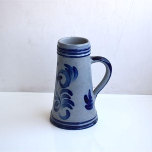 Puede incluir: Un jarro de cerveza de cerámica gris con asa azul y motivos florales azules. El jarro tiene un cuerpo cilíndrico que se estrecha hacia la base. Capacidad aproximada de 470 ml.