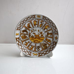 Può includere: Un piatto in ceramica bianca con un design marrone e oro che presenta gli animali dello zodiaco cinese. Il piatto ha un bordo decorativo con un motivo ripetitivo.
