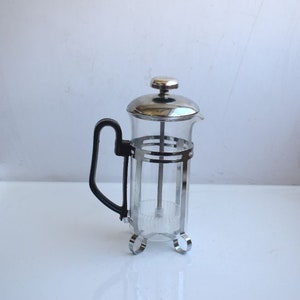 Op de afbeelding: Een zilveren en zwarte French press koffiezetapparaat met een glazen kan en een zwarte handgreep.