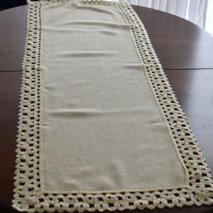 May include: A cream-colored table runner with a crocheted lace border. The runner is on a dark brown wooden table.