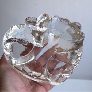 Könnte beinhalten: Eine klare Glasschale in Herzform mit mehreren kleineren herzförmigen Vertiefungen. Die Schale ist perfekt für die Aufbewahrung von kleinen Gegenständen wie Schmuck oder Nippes.
