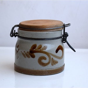 Puede incluir: Un tarro de cerámica blanca con un diseño floral marrón y una tapa de madera con un cierre de metal.