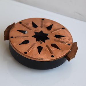 Peut inclure: Un brûleur d'encens en métal de couleur cuivre avec une découpe en forme d'étoile au centre et des découpes triangulaires sur le bord. Le brûleur a une base noire et des poignées en bois de chaque côté. Le design est circulaire.
